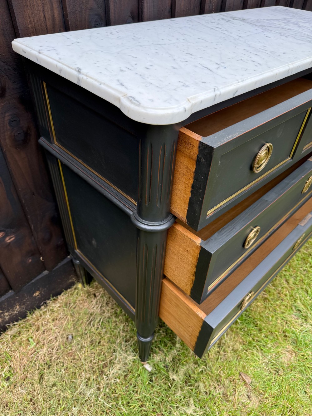 An early 20th-century painted Louis XVI style commode with a white marble top, dark green painted finish, gilded trim details, and cast brass ring pulls and keyhole escutcheons. View IV