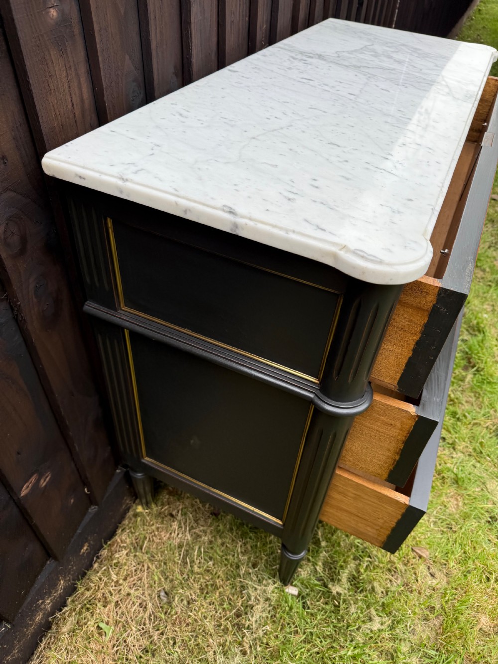 An early 20th-century painted Louis XVI style commode with a white marble top, dark green painted finish, gilded trim details, and cast brass ring pulls and keyhole escutcheons. View V