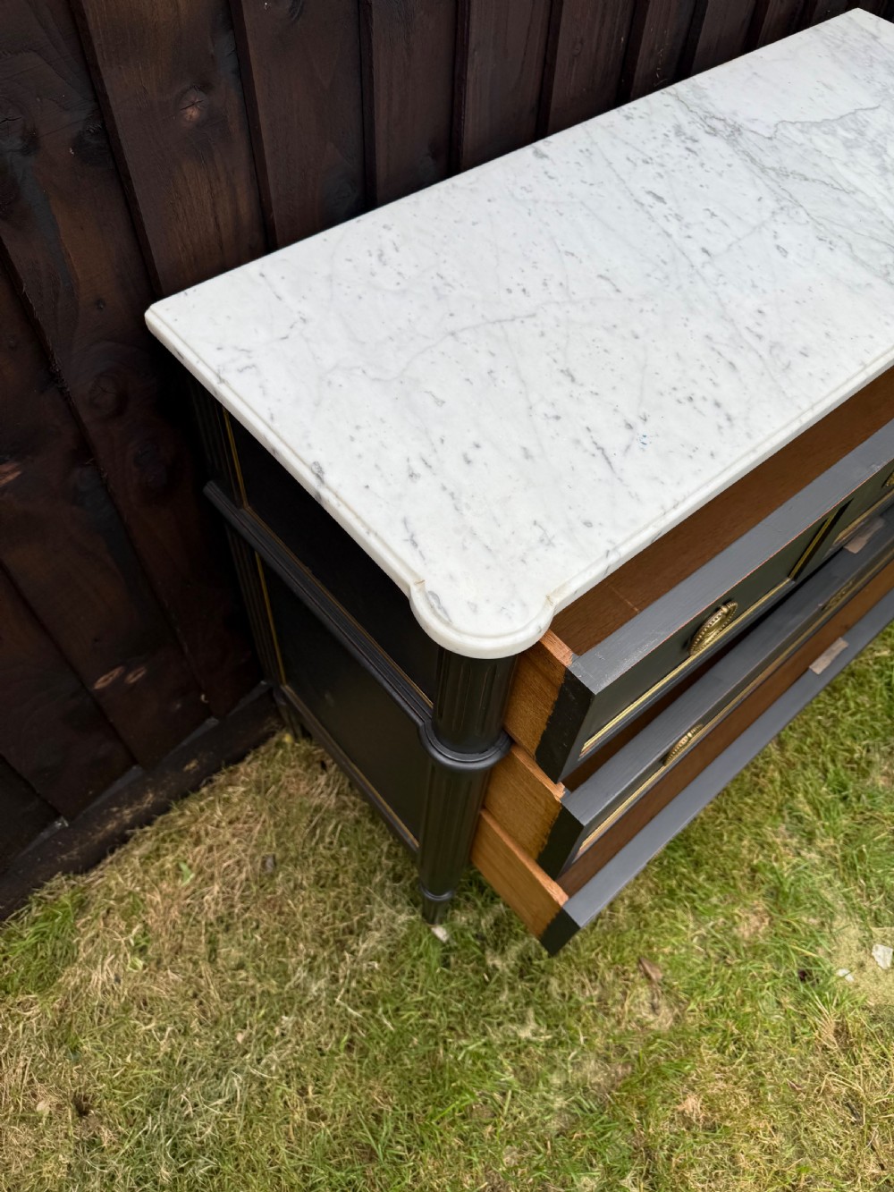 An early 20th-century painted Louis XVI style commode with a white marble top, dark green painted finish, gilded trim details, and cast brass ring pulls and keyhole escutcheons. View VI