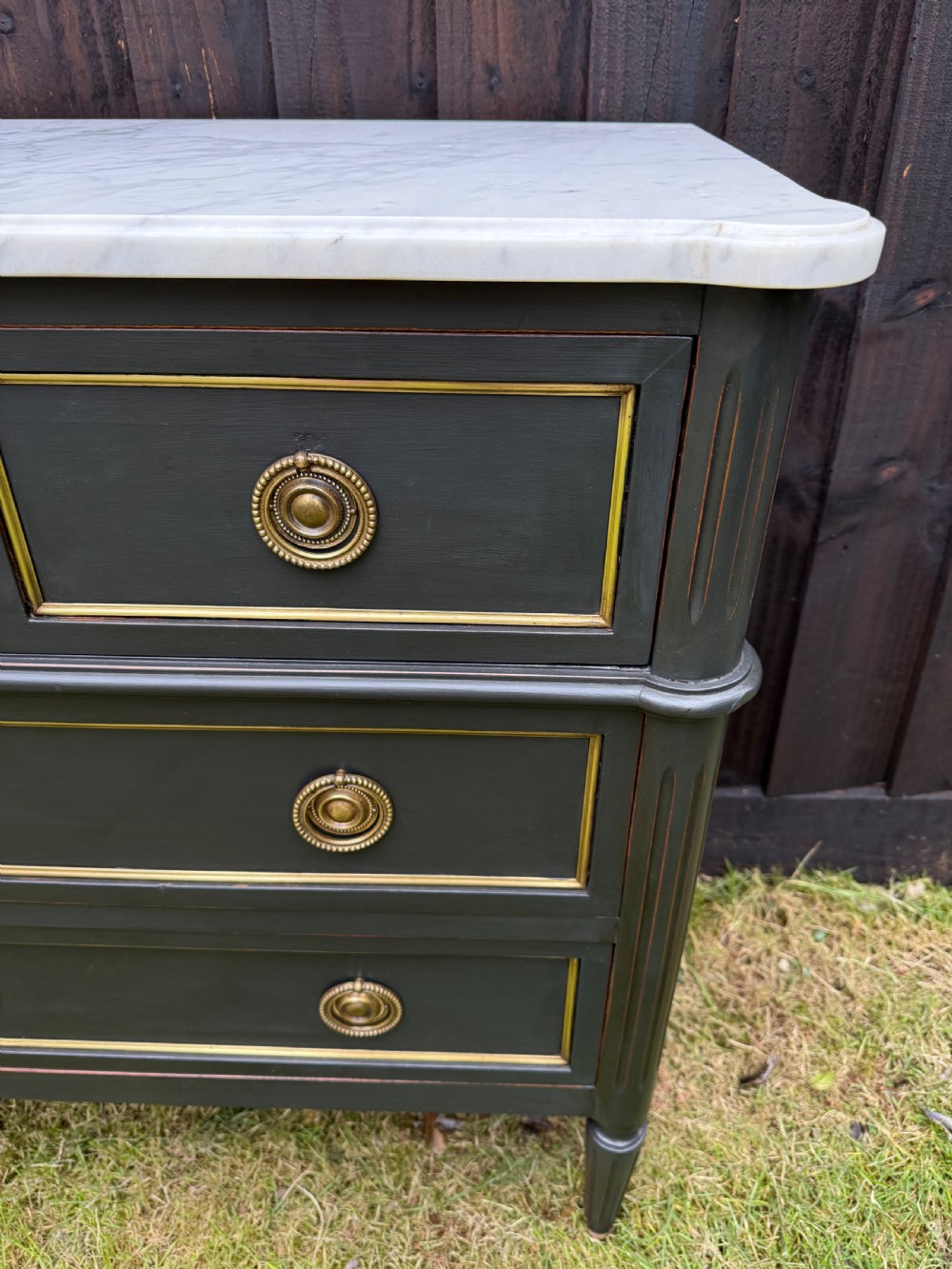 An early 20th-century painted Louis XVI style commode with a white marble top, dark green painted finish, gilded trim details, and cast brass ring pulls and keyhole escutcheons. View VII