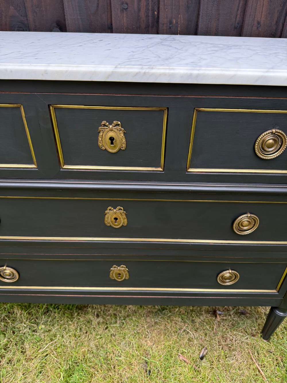 An early 20th-century painted Louis XVI style commode with a white marble top, dark green painted finish, gilded trim details, and cast brass ring pulls and keyhole escutcheons. View VIII