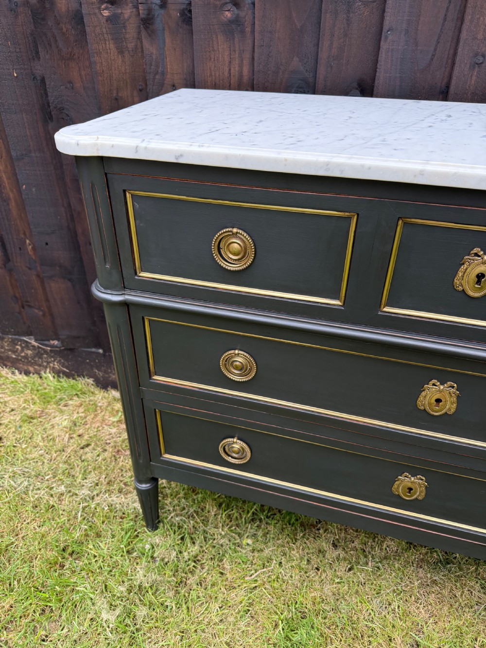 An early 20th-century painted Louis XVI style commode with a white marble top, dark green painted finish, gilded trim details, and cast brass ring pulls and keyhole escutcheons. View IX