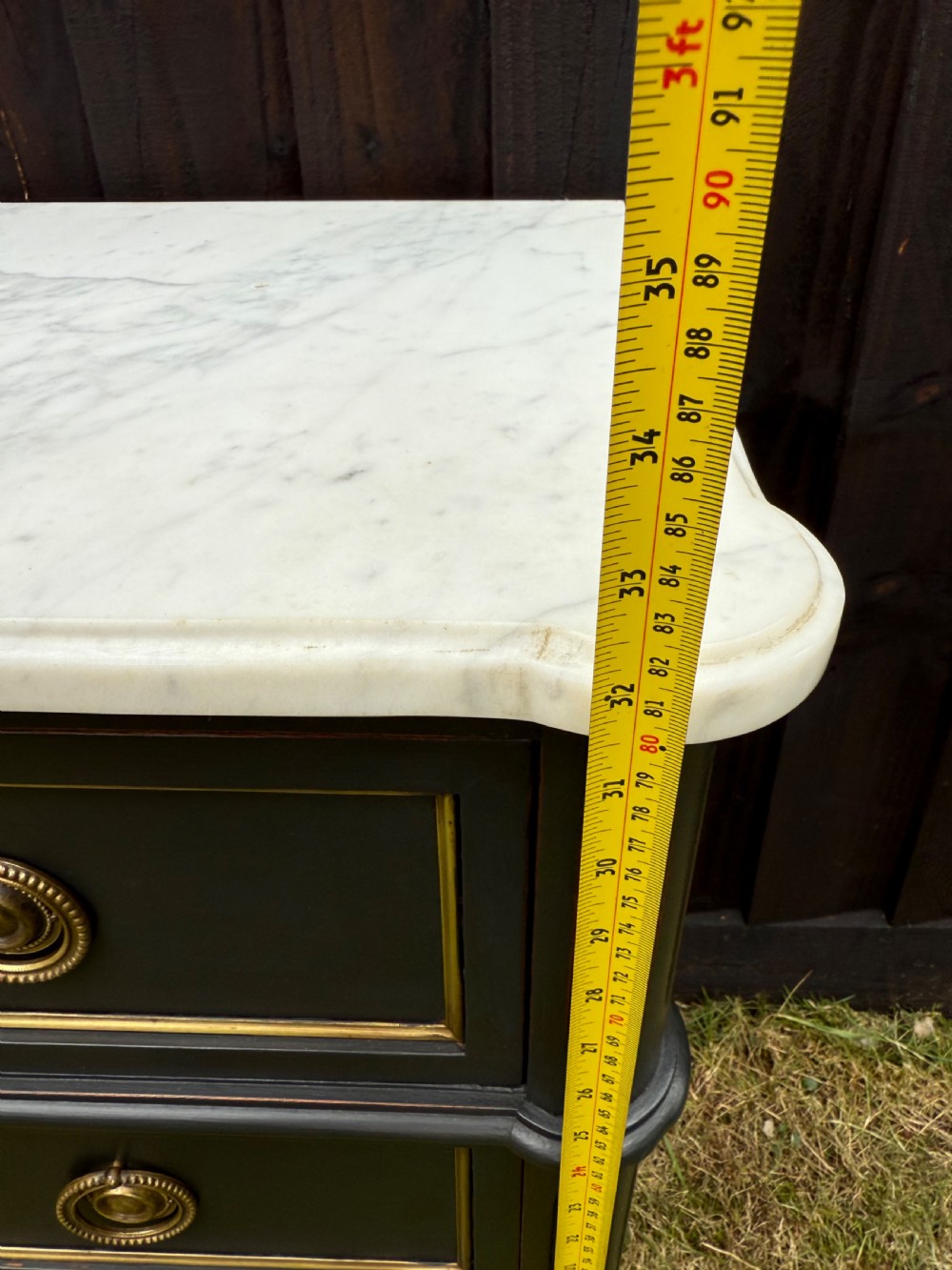 An early 20th-century painted Louis XVI style commode with a white marble top, dark green painted finish, gilded trim details, and cast brass ring pulls and keyhole escutcheons. View XII