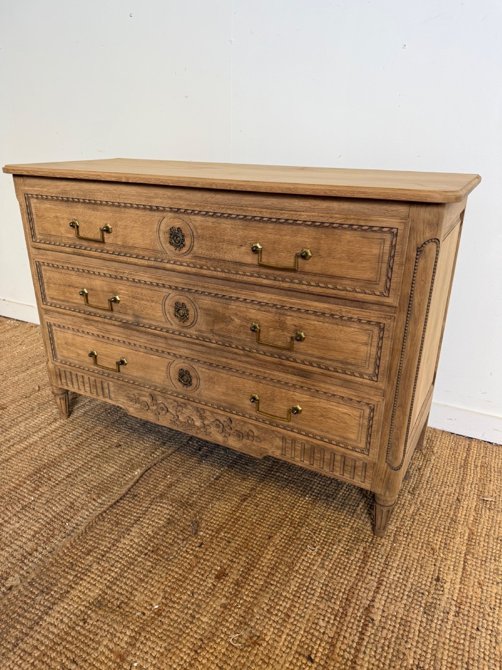 A rectangular scrubbed oak commode featuring three drawers with brass handles and subtle classical carved detailing on the facade.