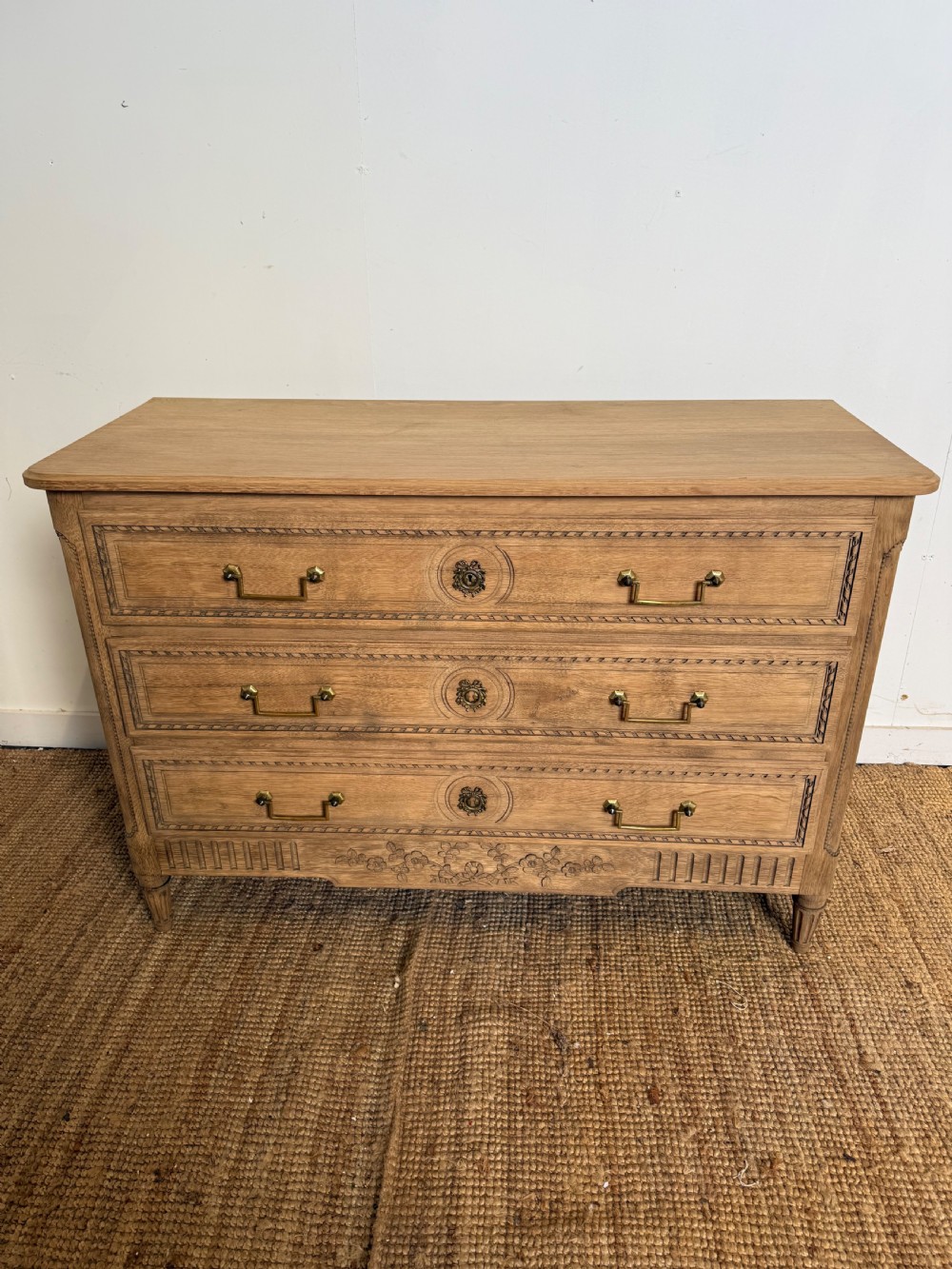 A rectangular scrubbed oak commode featuring three drawers with brass handles and subtle classical carved detailing on the facade. View III