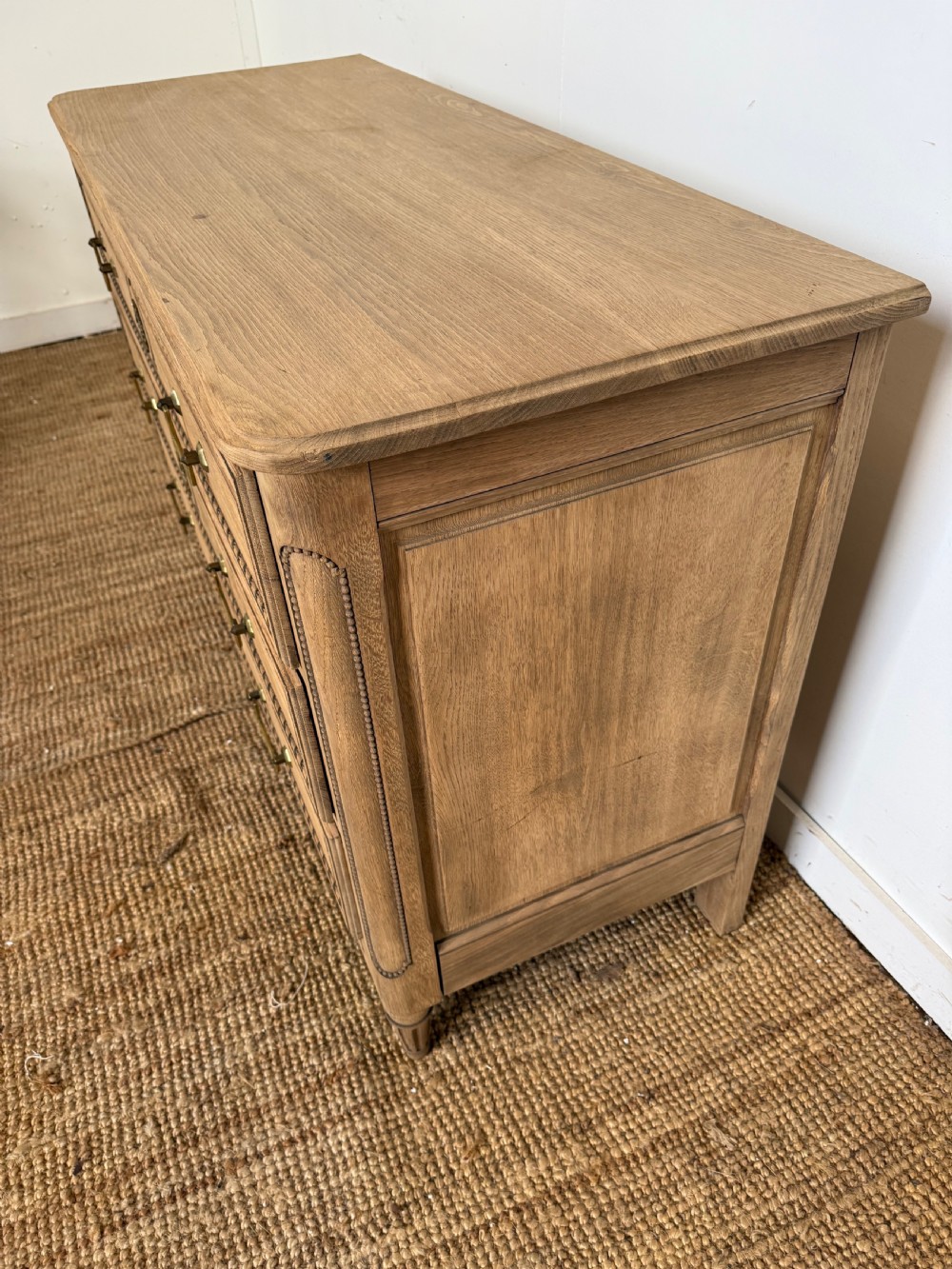A rectangular scrubbed oak commode featuring three drawers with brass handles and subtle classical carved detailing on the facade. View V