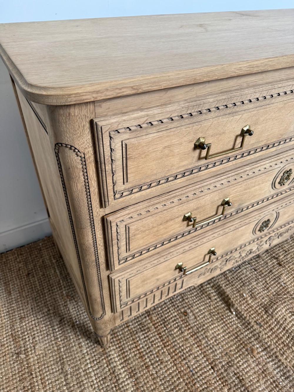 A rectangular scrubbed oak commode featuring three drawers with brass handles and subtle classical carved detailing on the facade. View VIII