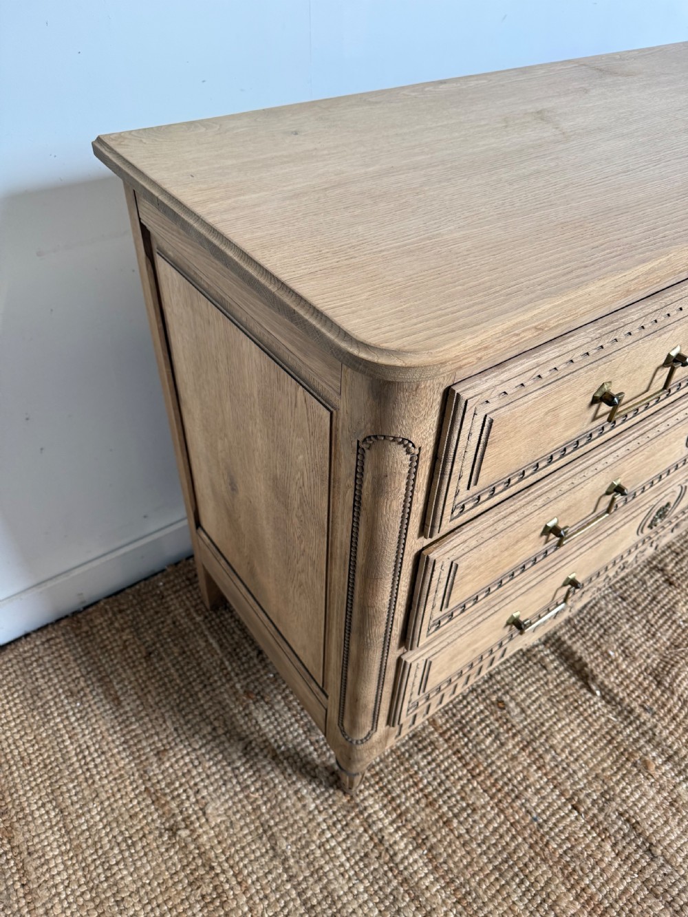 A rectangular scrubbed oak commode featuring three drawers with brass handles and subtle classical carved detailing on the facade. View IX