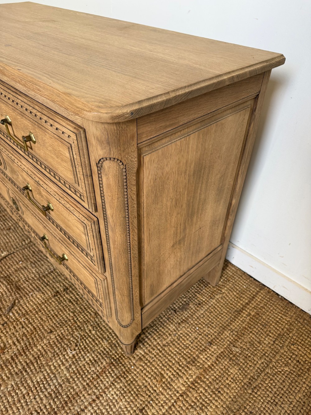 A rectangular scrubbed oak commode featuring three drawers with brass handles and subtle classical carved detailing on the facade. View X