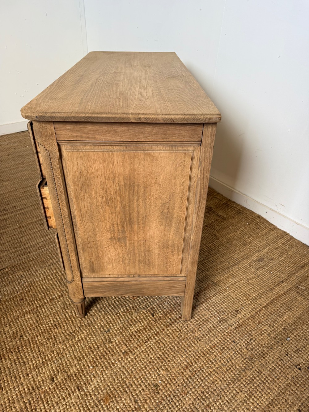 A rectangular scrubbed oak commode featuring three drawers with brass handles and subtle classical carved detailing on the facade. View XIII