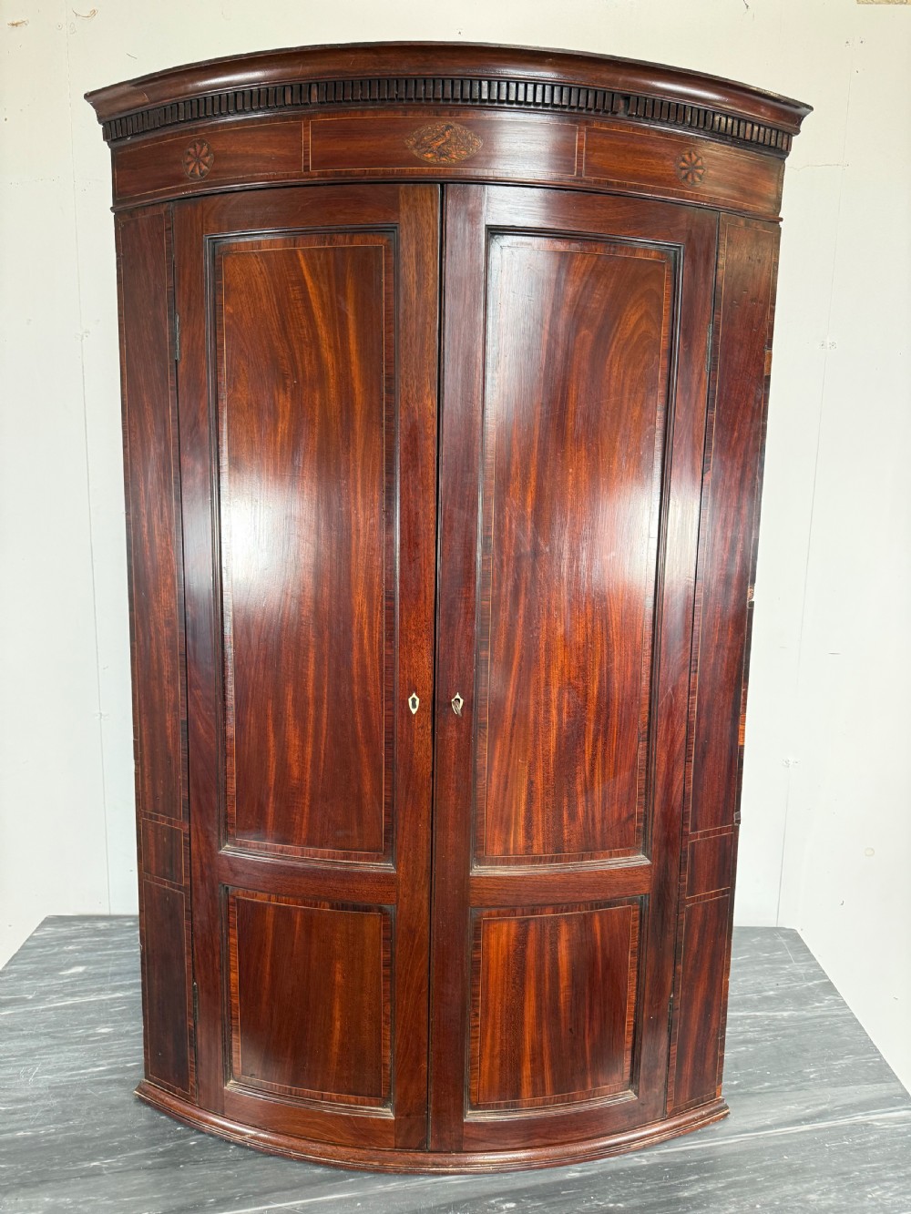 A Georgian mahogany bow-fronted corner cupboard featuring curved twin panelled doors, dentil moulding, and central marquetry inlay.