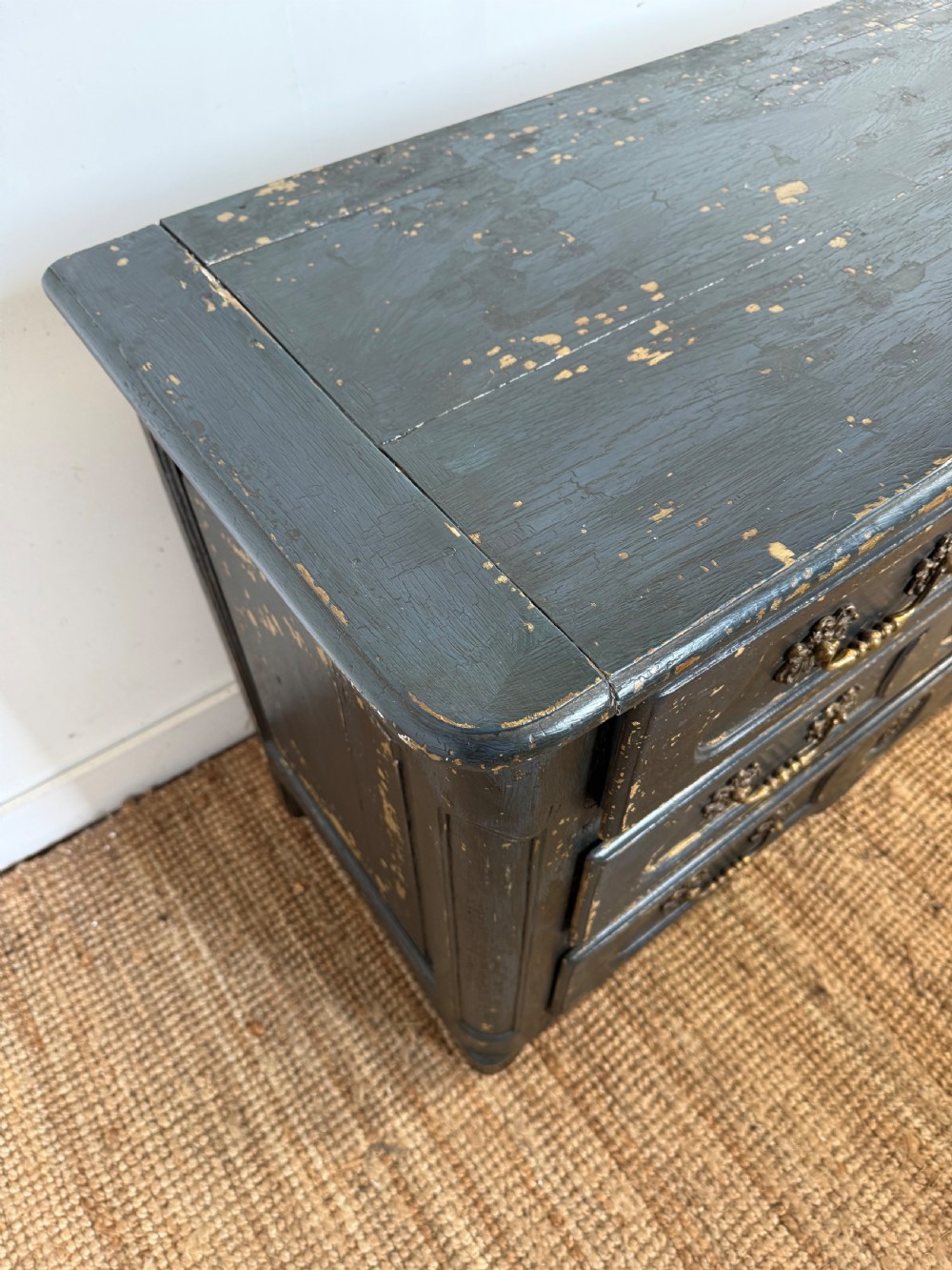 An antique dark-painted French commode featuring three rows of drawers with distressed black paint revealing bare wood underneath and decorative brass hardware. View V