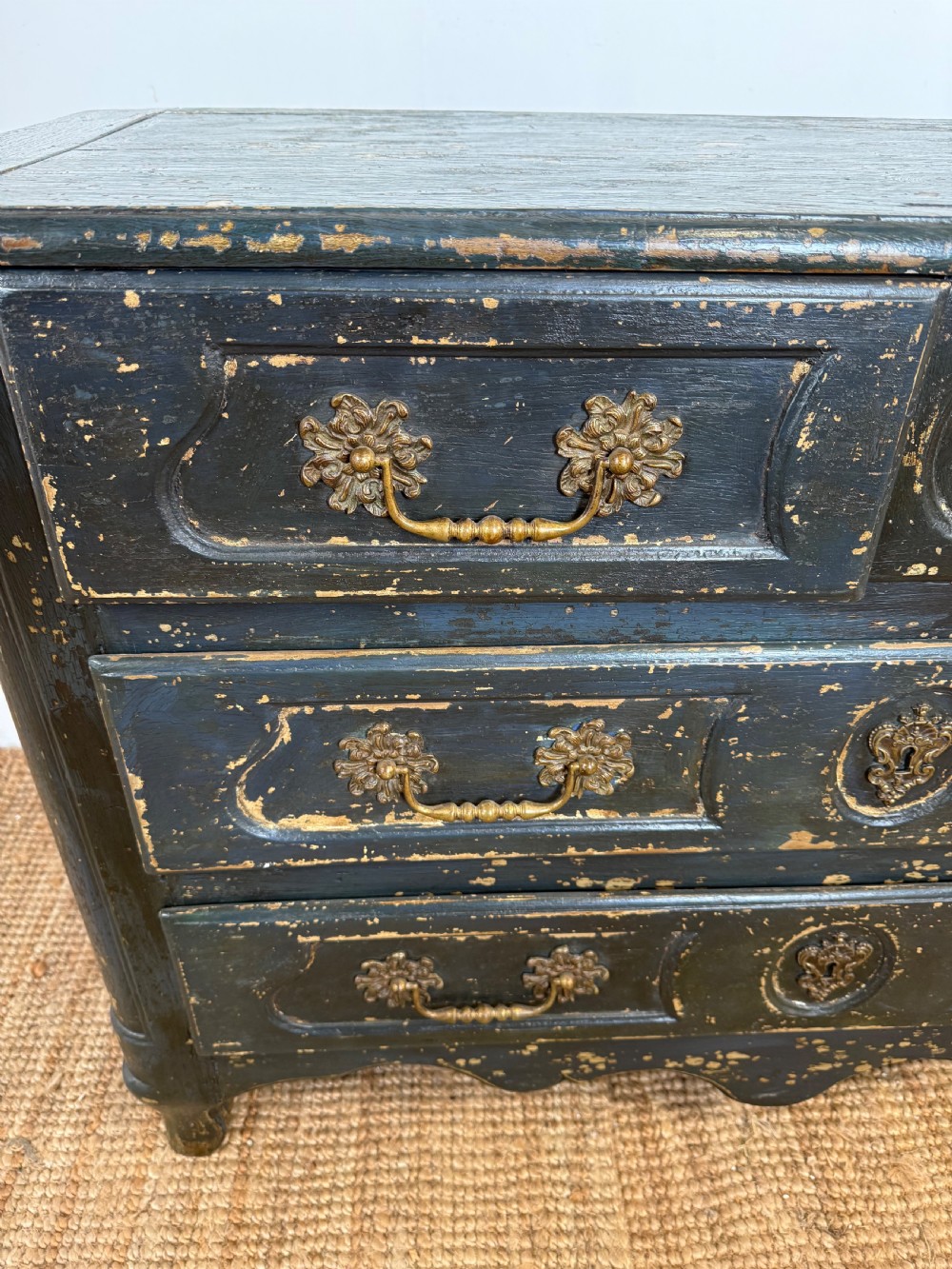 An antique dark-painted French commode featuring three rows of drawers with distressed black paint revealing bare wood underneath and decorative brass hardware. View VI