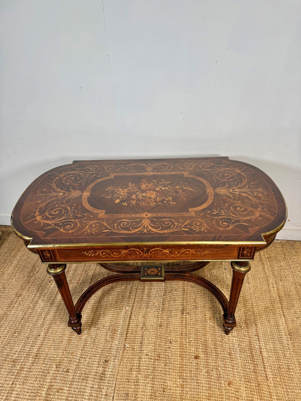 A French Napoleon III period centre table featuring floral wood marquetry, brass edges, tapered fluted legs, and an arched lower stretcher.