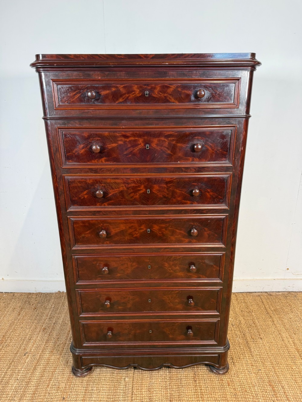 A tall, narrow flame mahogany semainier chest featuring seven drawers with turned knobs and central keyholes set on a moulded plinth base.