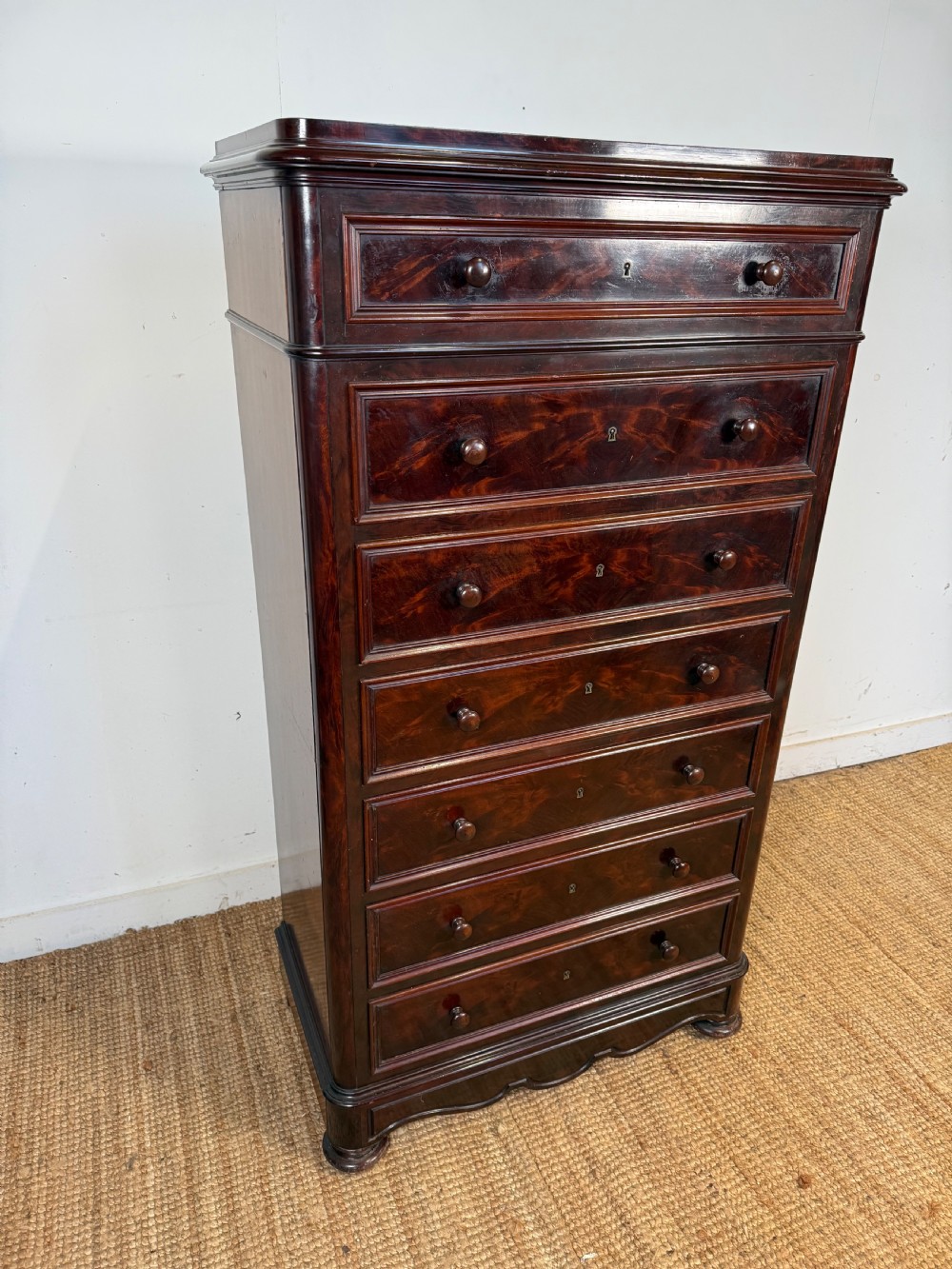 A tall, narrow flame mahogany semainier chest featuring seven drawers with turned knobs and central keyholes set on a moulded plinth base. View II