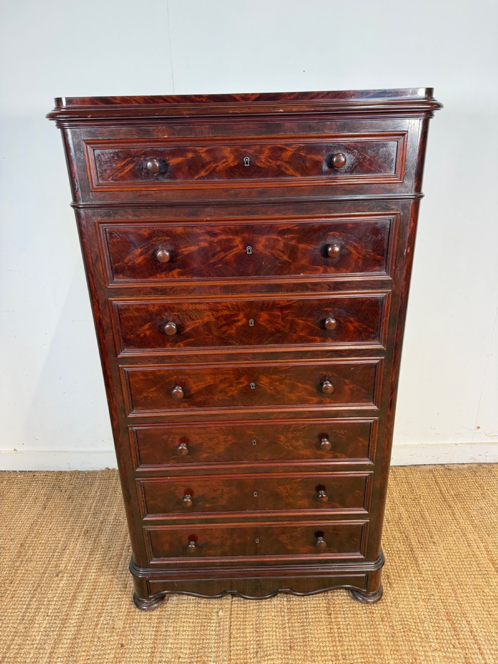 A tall, narrow flame mahogany semainier chest featuring seven drawers with turned knobs and central keyholes set on a moulded plinth base. View III