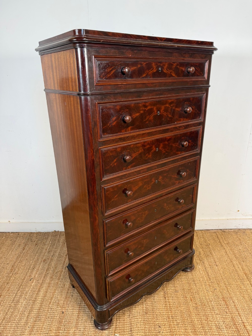 A tall, narrow flame mahogany semainier chest featuring seven drawers with turned knobs and central keyholes set on a moulded plinth base. View V