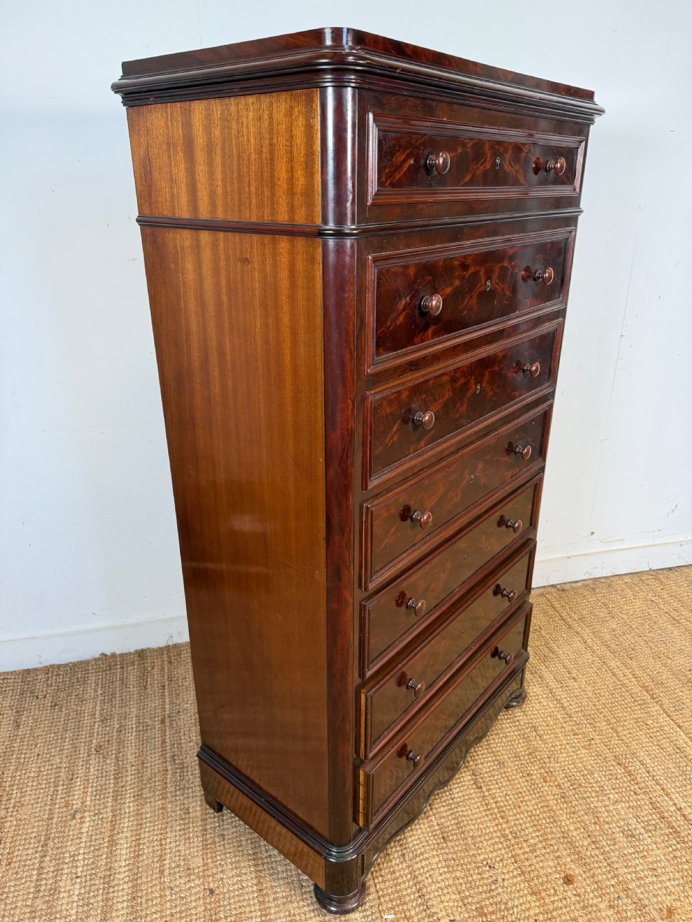 A tall, narrow flame mahogany semainier chest featuring seven drawers with turned knobs and central keyholes set on a moulded plinth base. View VI