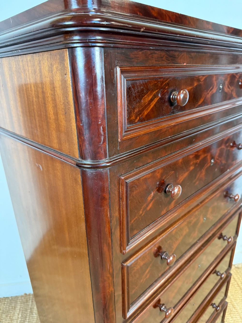 A tall, narrow flame mahogany semainier chest featuring seven drawers with turned knobs and central keyholes set on a moulded plinth base. View VII