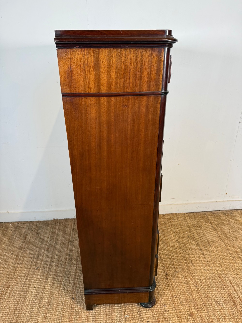 A tall, narrow flame mahogany semainier chest featuring seven drawers with turned knobs and central keyholes set on a moulded plinth base. View VIII