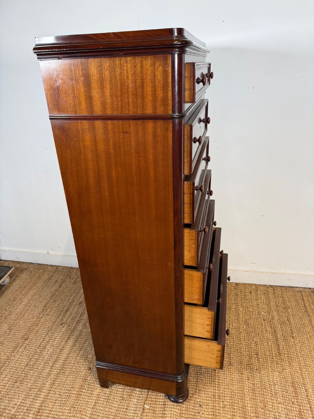 A tall, narrow flame mahogany semainier chest featuring seven drawers with turned knobs and central keyholes set on a moulded plinth base. View IX