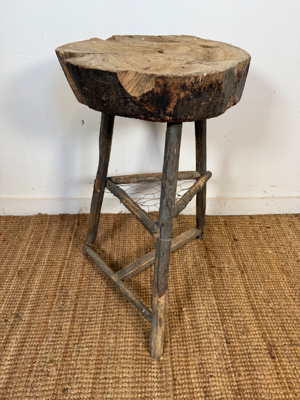 A rustic timber chopping block featuring a thick round log cross-section top supported by three weathered wooden legs with a wire mesh shelf.
