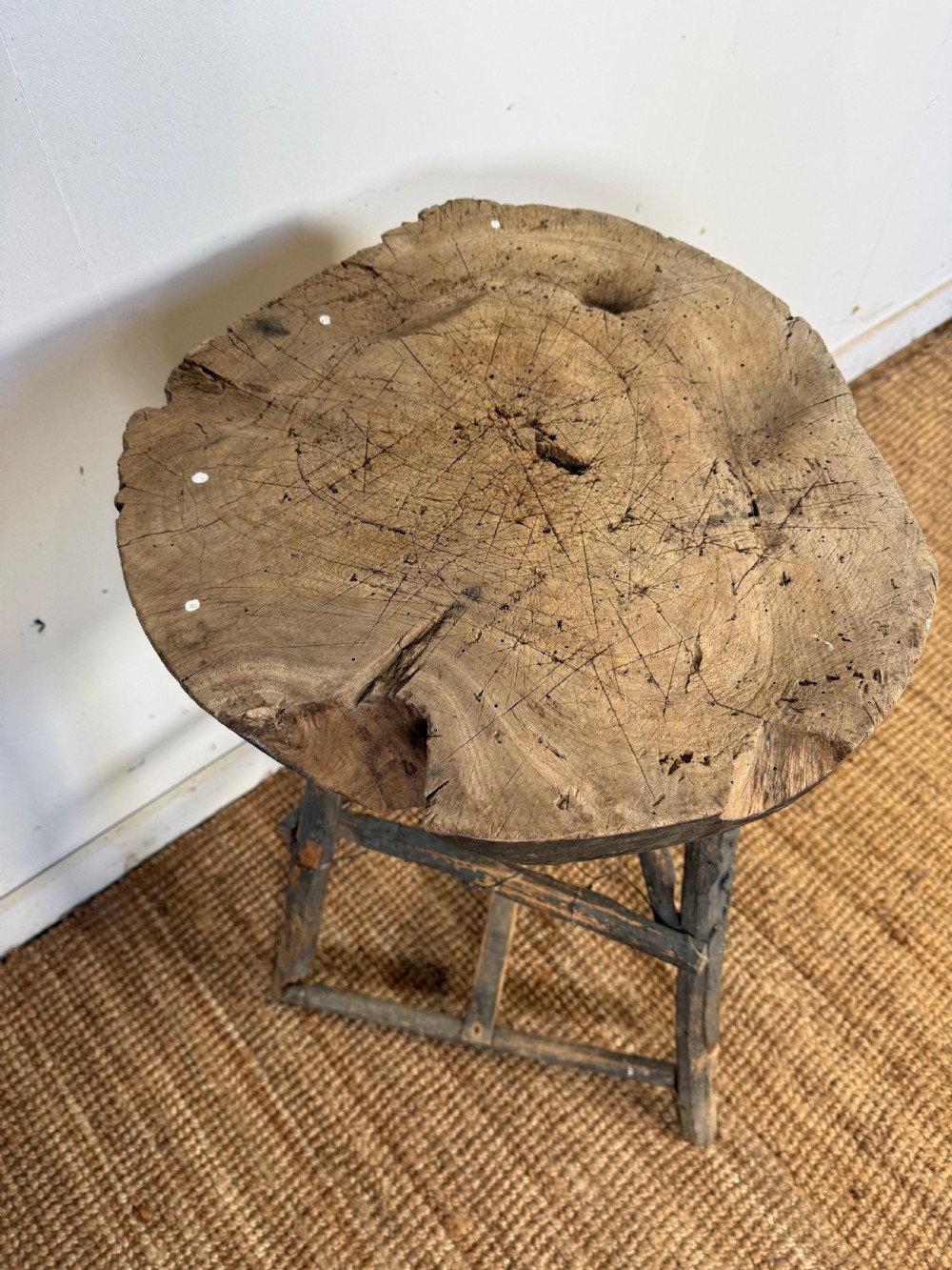 A rustic timber chopping block featuring a thick round log cross-section top supported by three weathered wooden legs with a wire mesh shelf. View IV