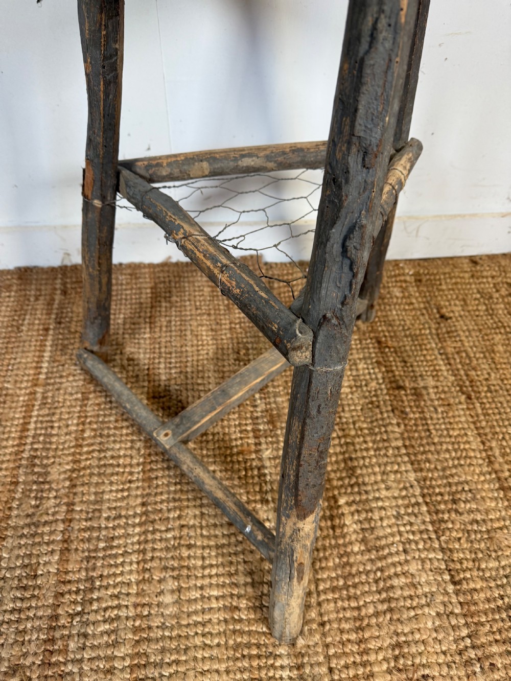 A rustic timber chopping block featuring a thick round log cross-section top supported by three weathered wooden legs with a wire mesh shelf. View V