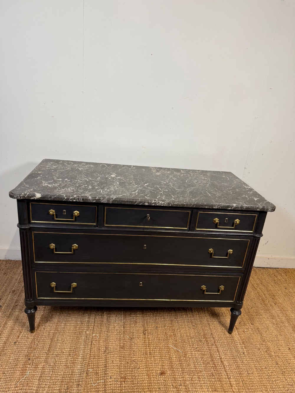 An early 19th-century black painted Louis XVI style commode featuring a grey marble top, brass trim, and fluted column legs.