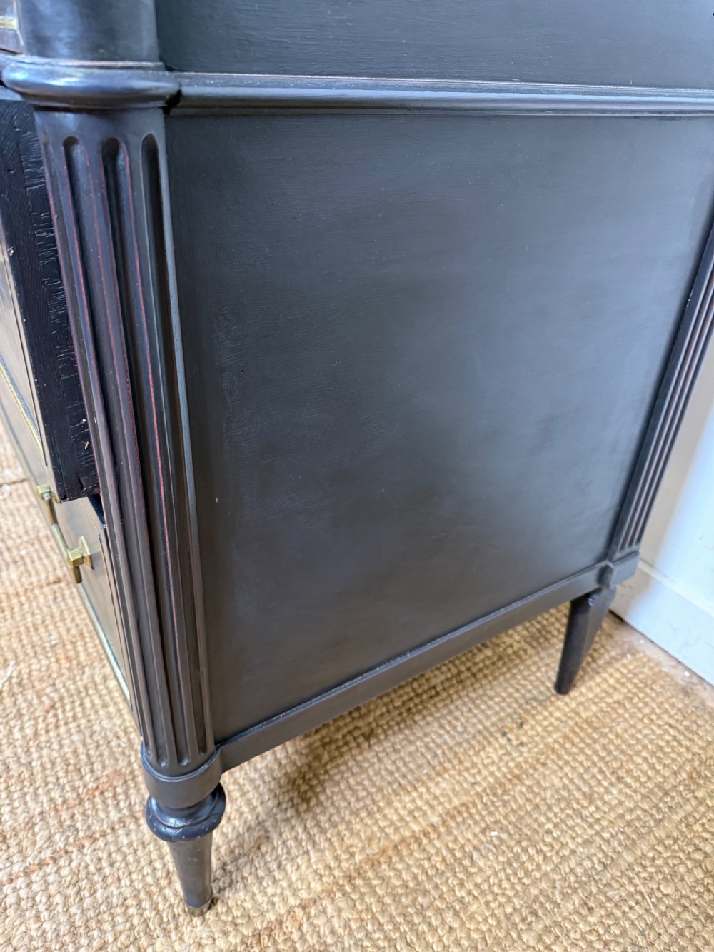 An early 19th-century black painted Louis XVI style commode featuring a grey marble top, brass trim, and fluted column legs. View XVI