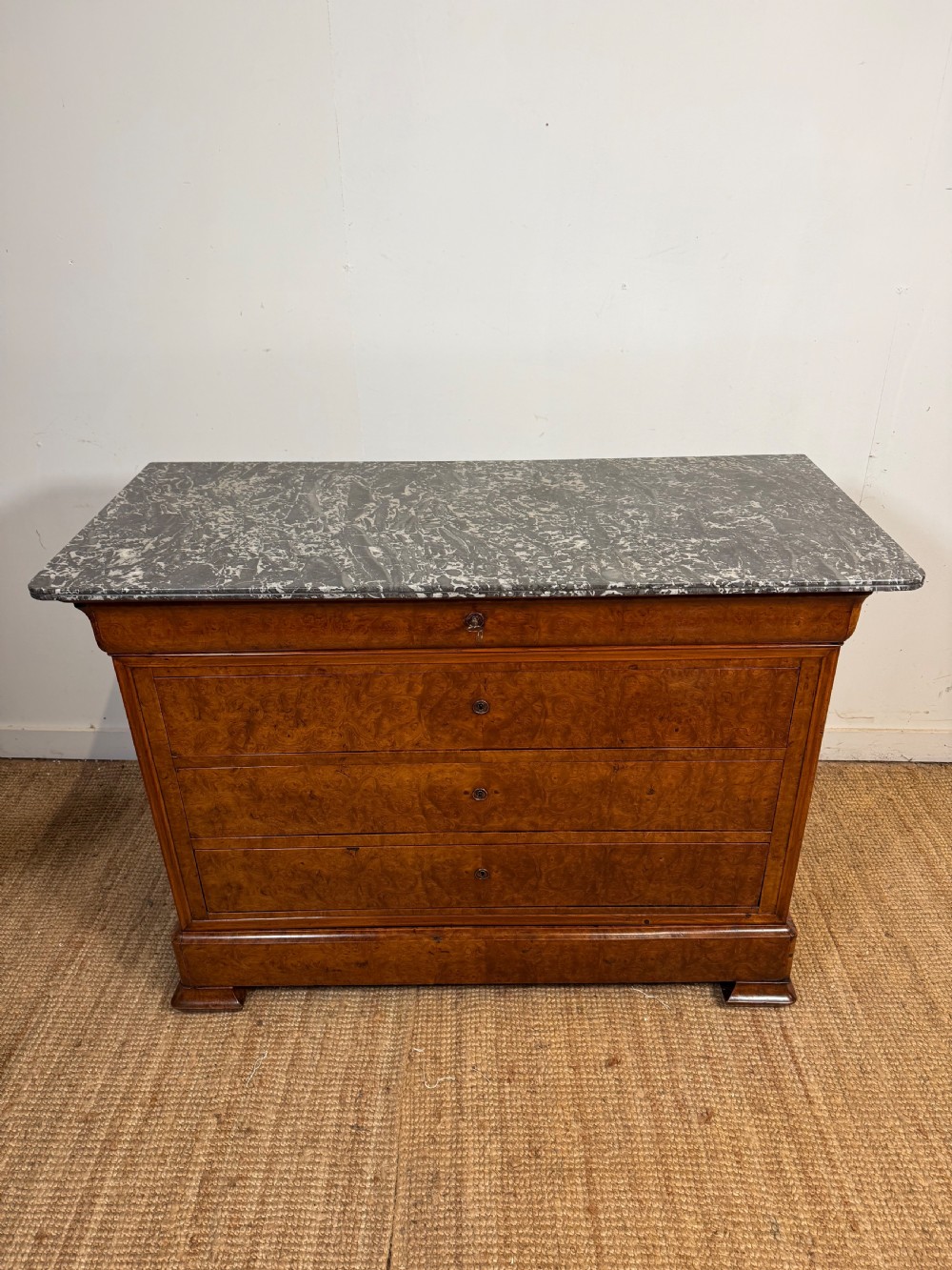 A rectangular 19th-century burr elm commode featuring a grey mottled marble top and three long drawers with central key escutcheons.