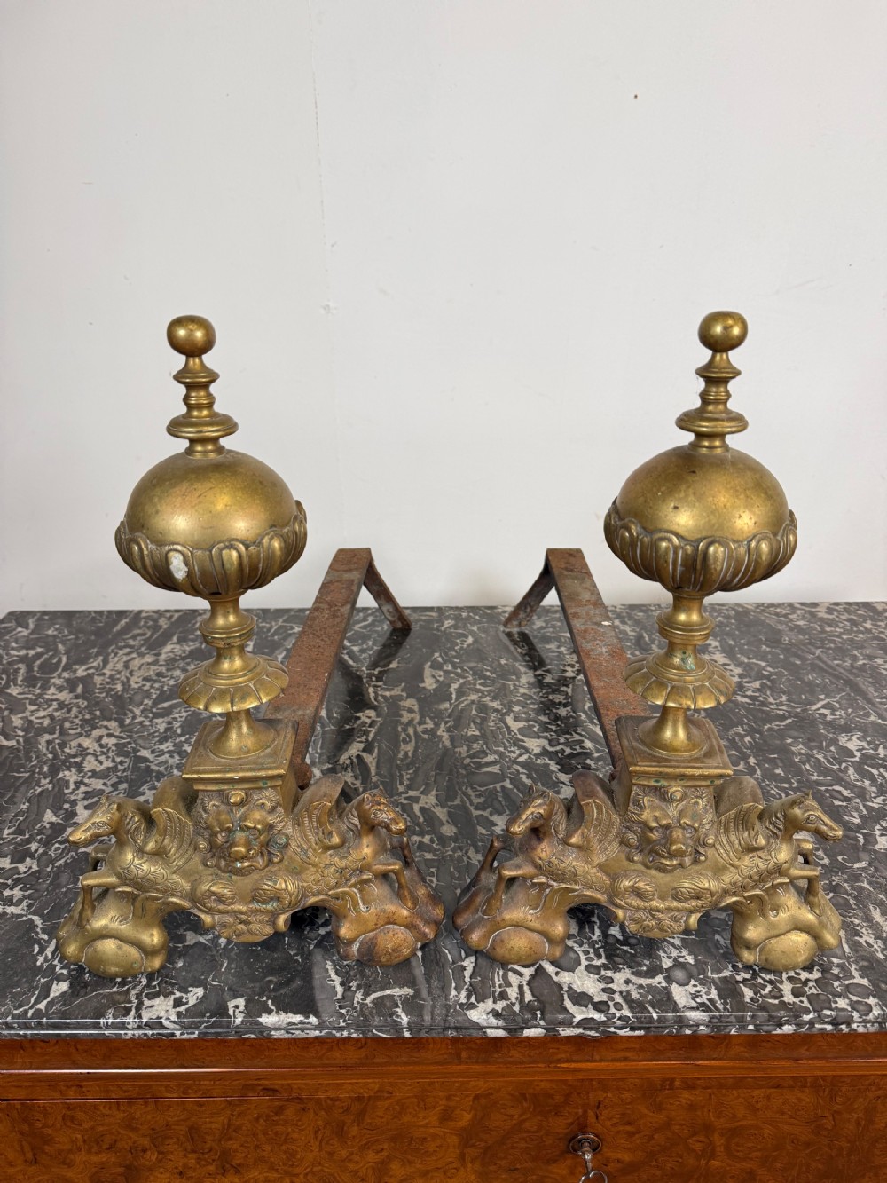 A pair of antique brass fire dogs featuring urn finials, gadrooned details, relief-carved creature bases, and iron billet bars.