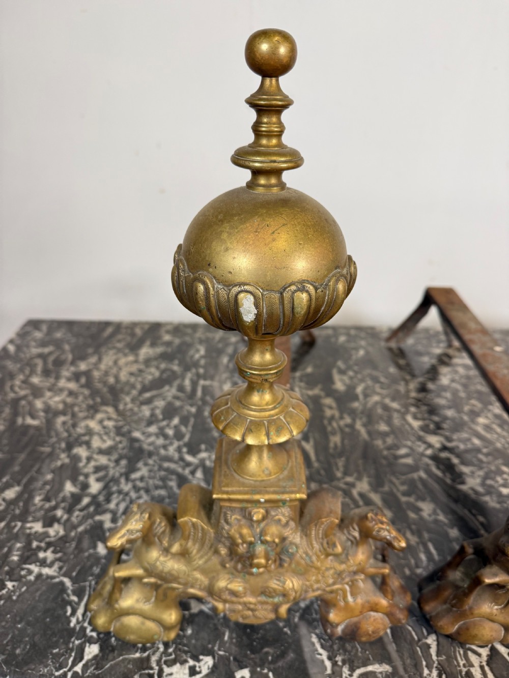 A pair of antique brass fire dogs featuring urn finials, gadrooned details, relief-carved creature bases, and iron billet bars. View II