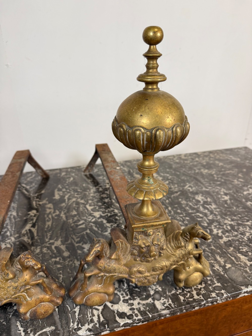 A pair of antique brass fire dogs featuring urn finials, gadrooned details, relief-carved creature bases, and iron billet bars. View III