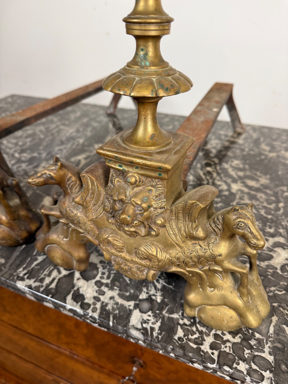 A pair of antique brass fire dogs featuring urn finials, gadrooned details, relief-carved creature bases, and iron billet bars. View V