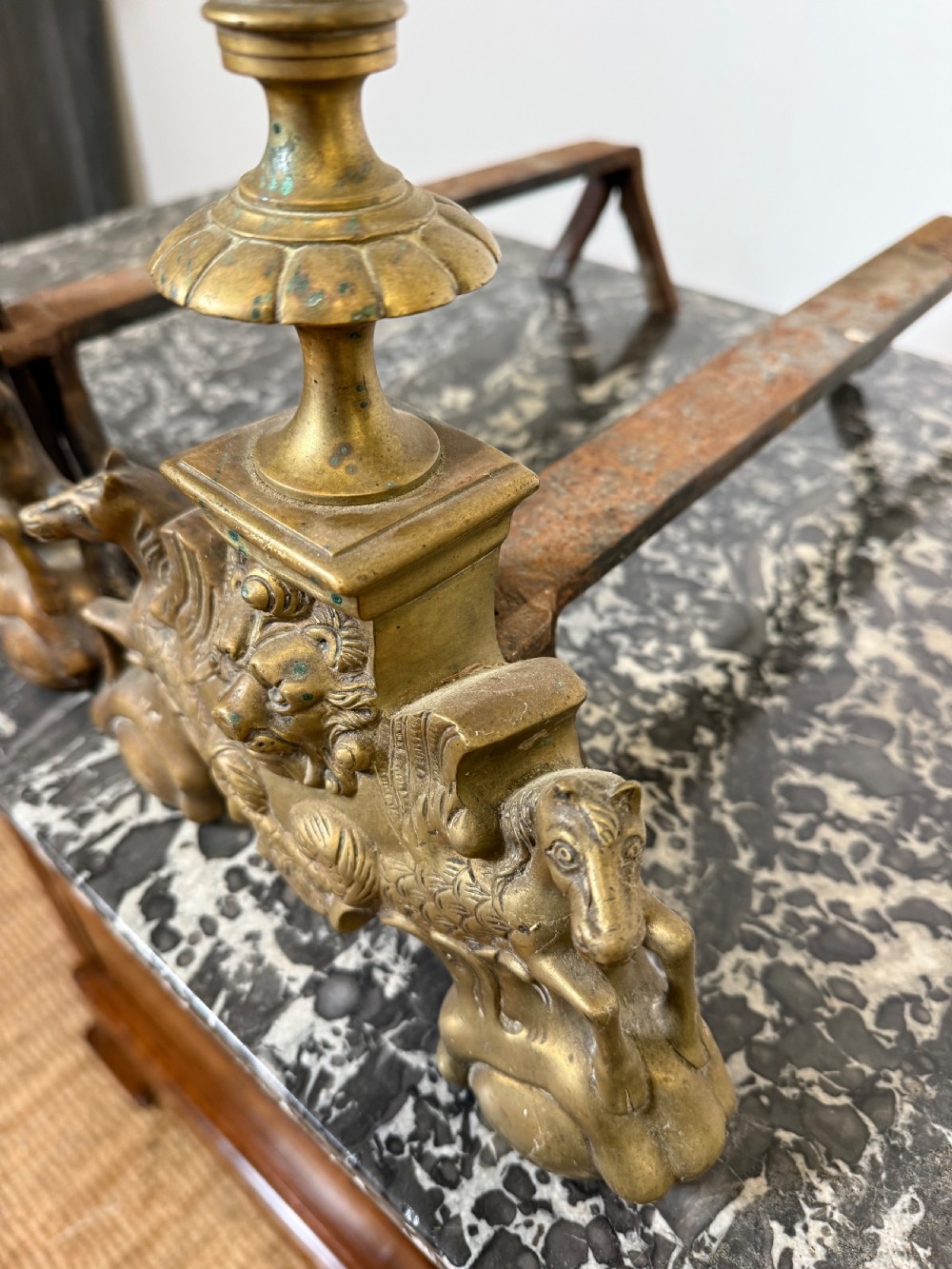 A pair of antique brass fire dogs featuring urn finials, gadrooned details, relief-carved creature bases, and iron billet bars. View VI