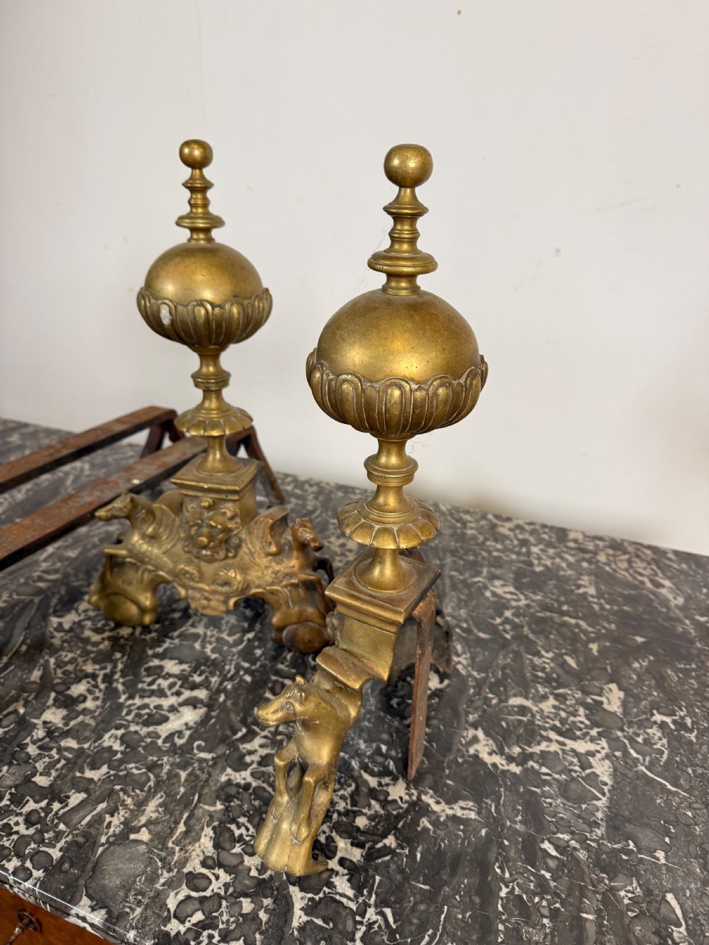 A pair of antique brass fire dogs featuring urn finials, gadrooned details, relief-carved creature bases, and iron billet bars. View IX