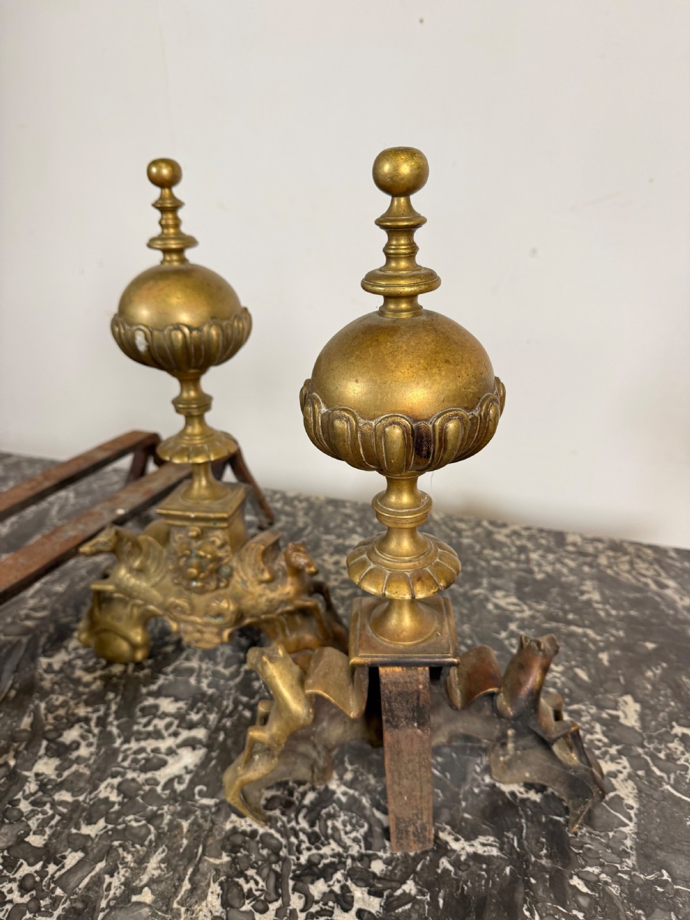 A pair of antique brass fire dogs featuring urn finials, gadrooned details, relief-carved creature bases, and iron billet bars. View X