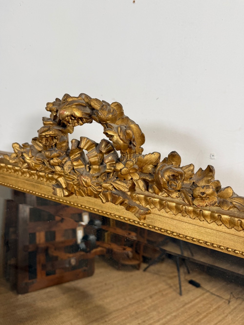 A rectangular antique wall mirror featuring a prominent ornate floral and ribbon carved crest in a gilded finish. View IV