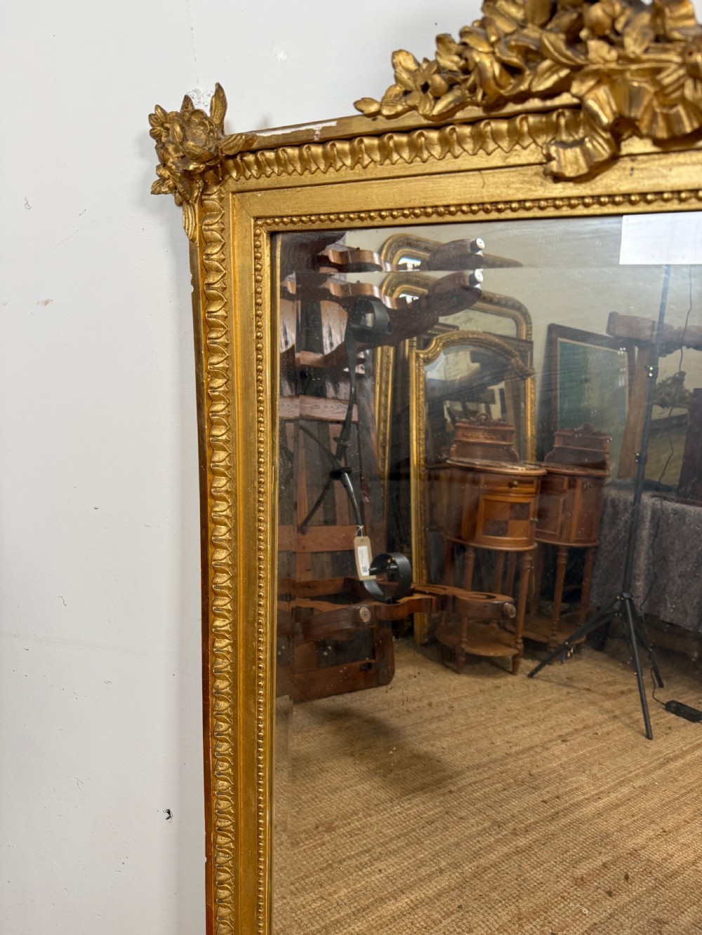 A rectangular antique wall mirror featuring a prominent ornate floral and ribbon carved crest in a gilded finish. View VII
