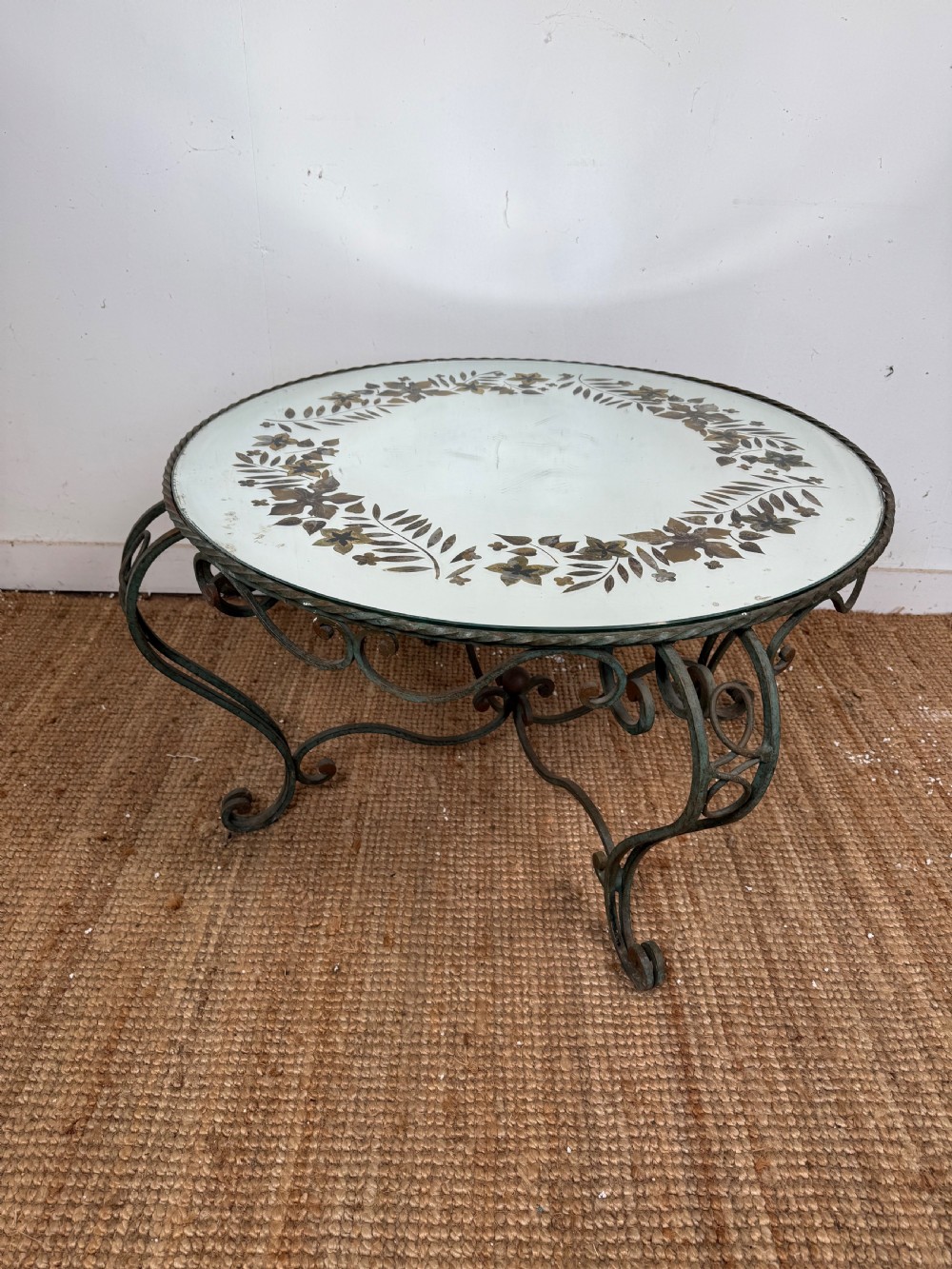 A round Art Deco coffee table featuring a verdigris wrought iron frame with scroll legs and a circular glass top decorated with a painted floral border.