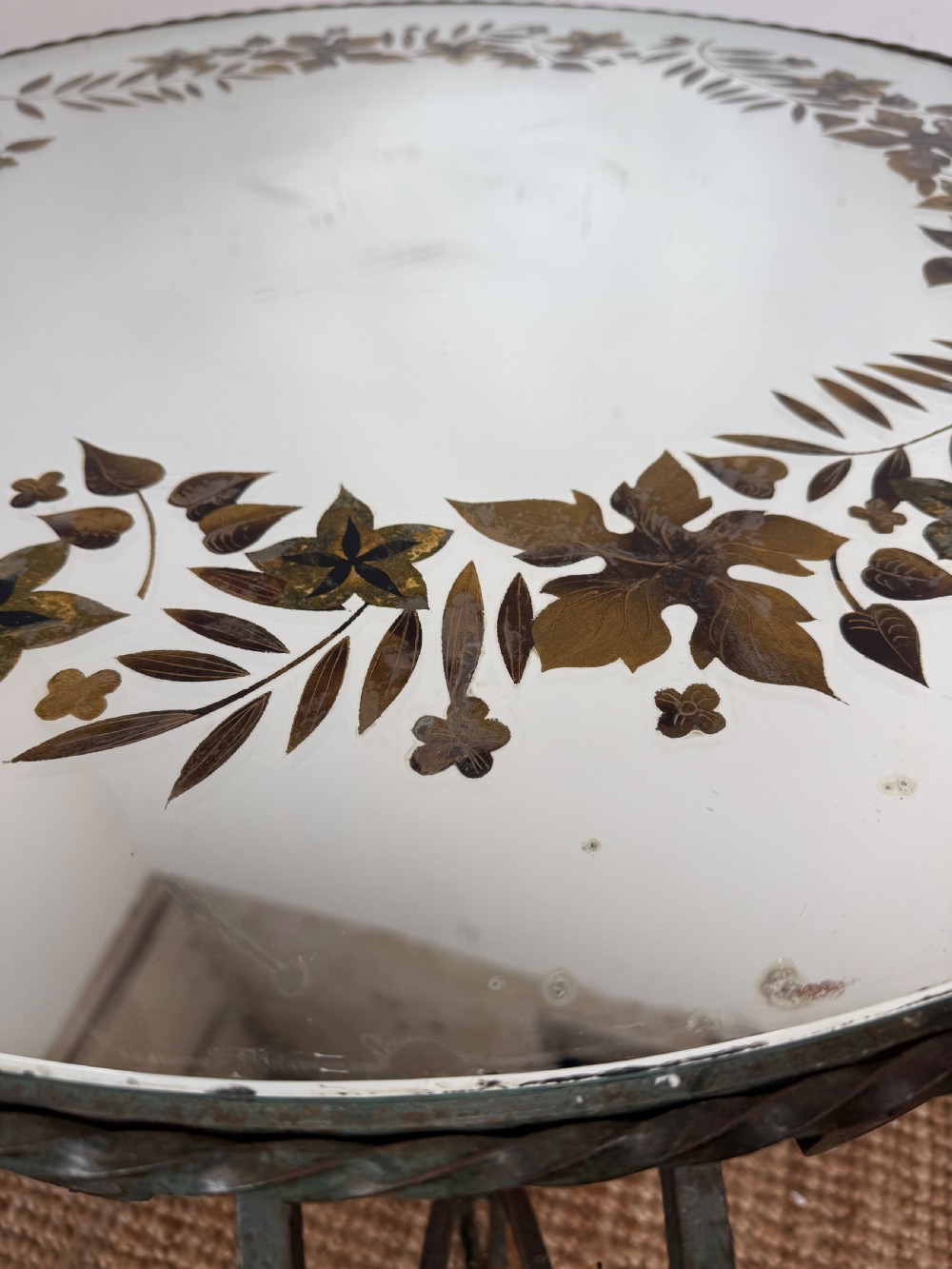 A round Art Deco coffee table featuring a verdigris wrought iron frame with scroll legs and a circular glass top decorated with a painted floral border. View V