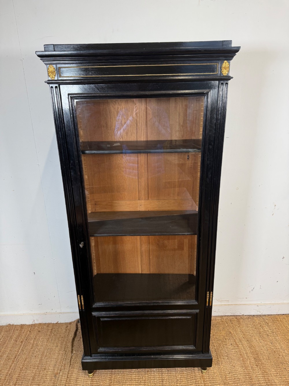 A narrow 19th-century Napoleon III ebonised wooden bookcase featuring a single glazed glass door, decorative gilt brass mounts, and a warm oak lined interior.