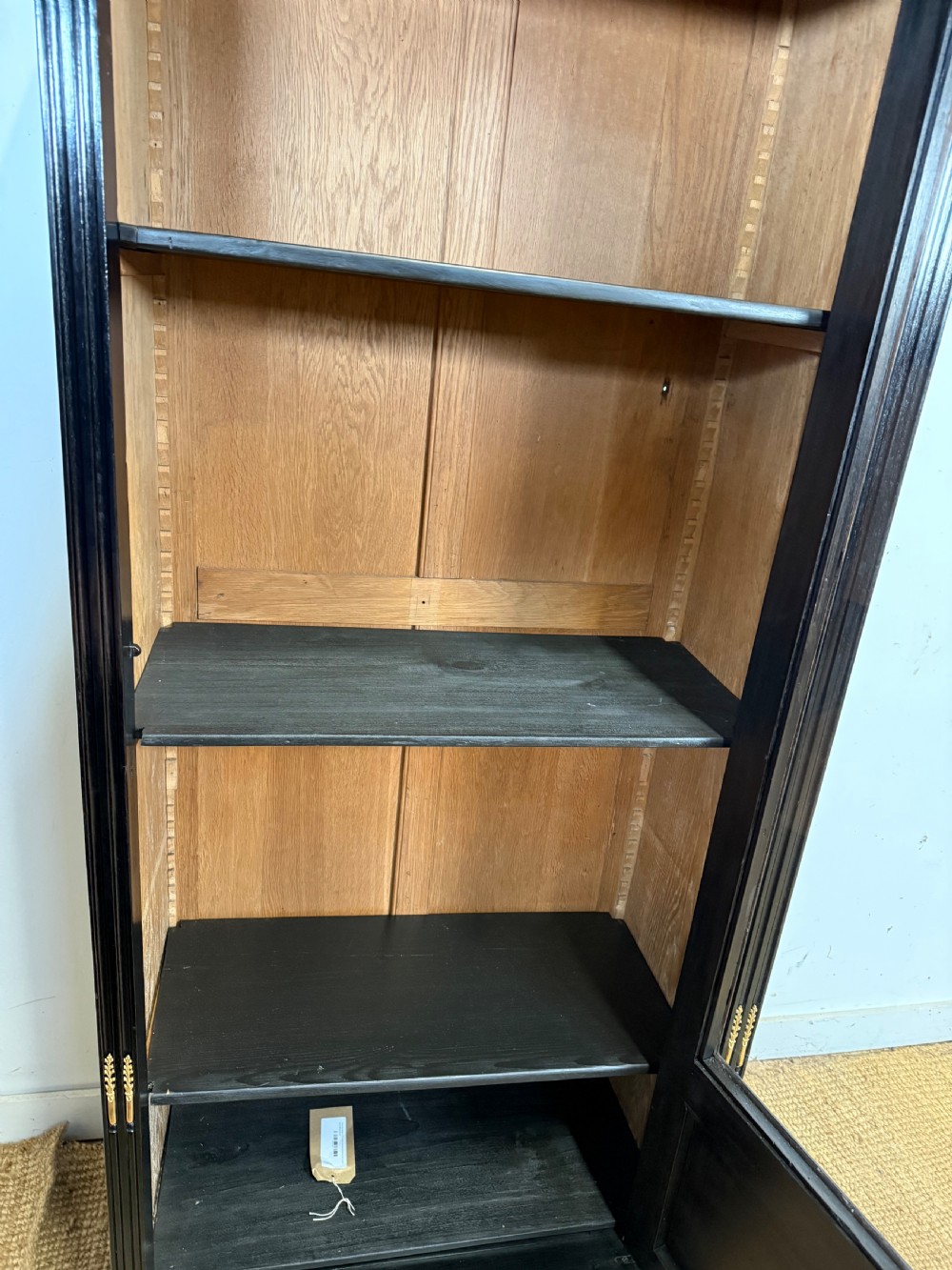 A narrow 19th-century Napoleon III ebonised wooden bookcase featuring a single glazed glass door, decorative gilt brass mounts, and a warm oak lined interior. View XII
