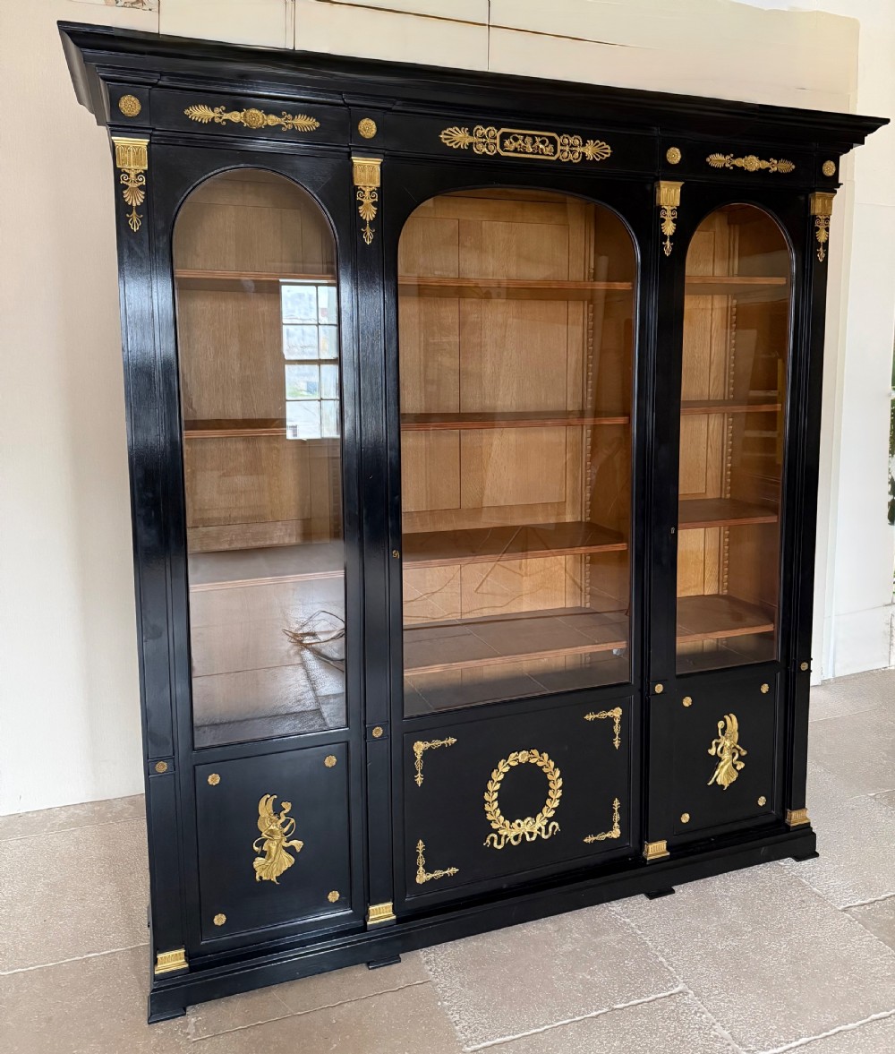 A large ebonised three-door glazed bookcase featuring gold-toned gilt-bronze mounts, arched glass panels, and an oak interior.