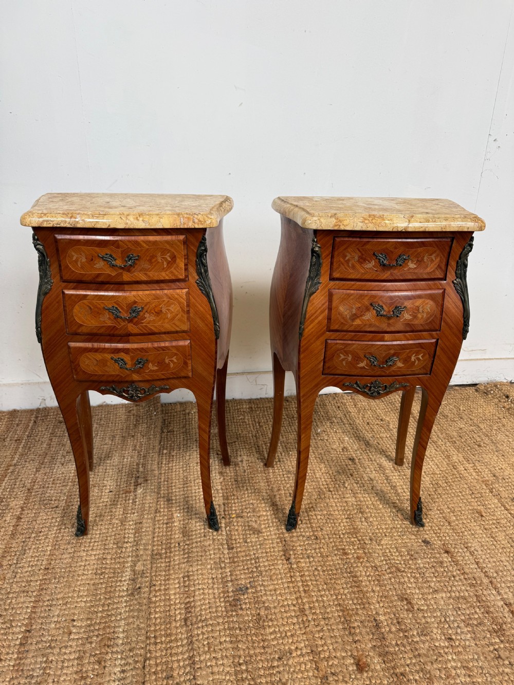 A pair of French Louis XV style bedside cabinets featuring variegated yellow marble tops, three marquetry inlaid drawers, brass mounts, and slender cabriole legs.