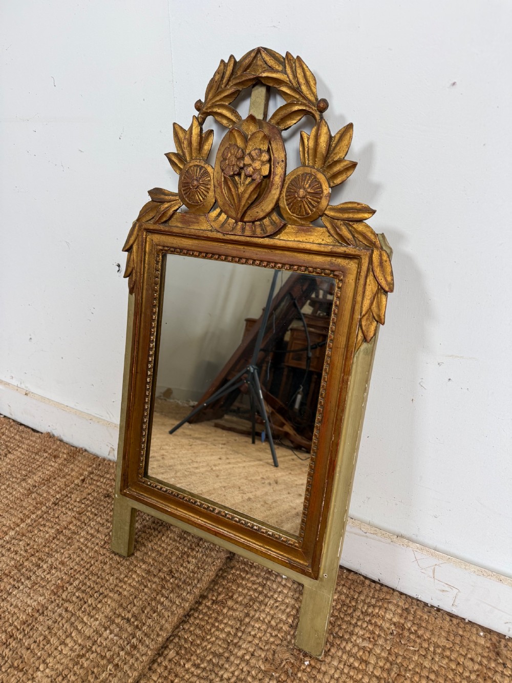 A compact Louis XVI style wall mirror featuring a gilded carved wood frame with beaded moulding and a prominent floral crest.