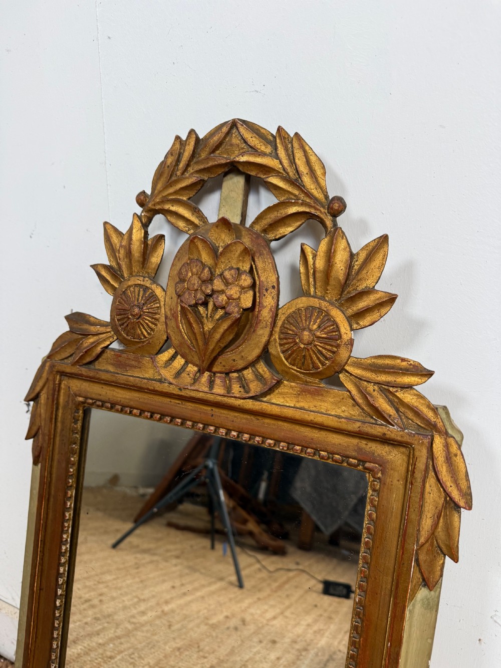 A compact Louis XVI style wall mirror featuring a gilded carved wood frame with beaded moulding and a prominent floral crest. View II