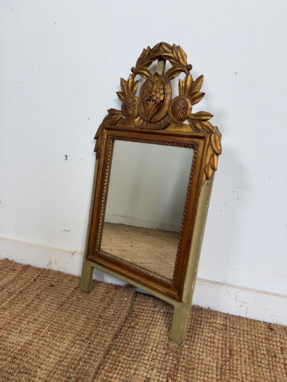 A compact Louis XVI style wall mirror featuring a gilded carved wood frame with beaded moulding and a prominent floral crest. View VII